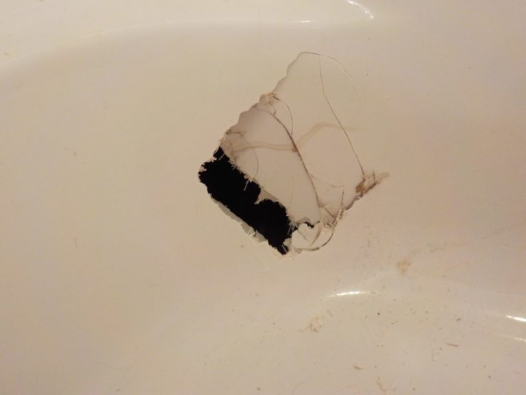 A close-up of a white bathtub surface with a large, irregular hole showing a dark area beneath and cracks spreading from the edges. There are some faint dirt marks around the damaged area.