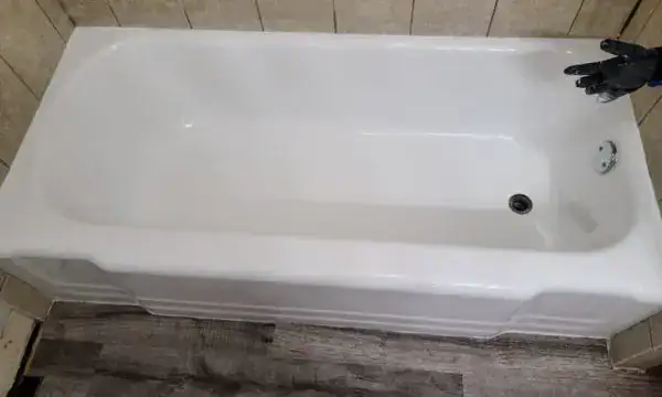 A clean, white bathtub with beige tiled walls and dark wood-patterned flooring. A black-gloved hand is visible near the top right corner of the image.